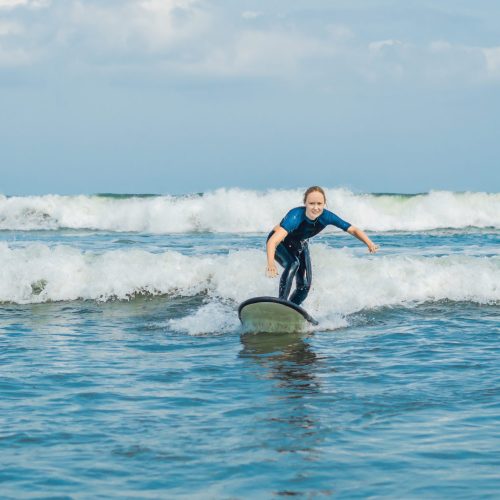 Joyful young woman beginner surfer with blue surf has fun on small sea waves. Active family lifestyle, people outdoor water sport lesson and swimming activity on surf camp summer vacation.